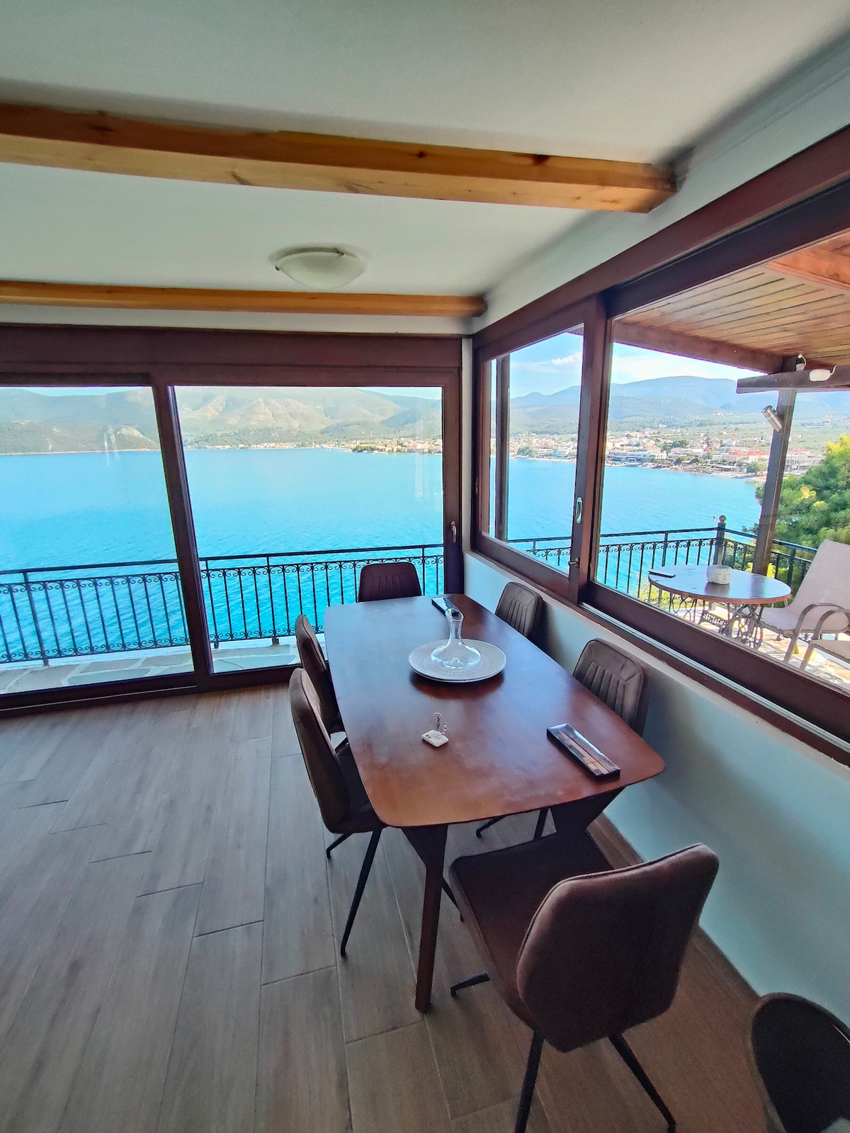 A modern dining area features a rectangular wooden table surrounded by six dining chairs. Large windows provide expansive views of the sea and mountains, allowing natural light to fill the space. The serene setting invites guests to enjoy meals while admiring the picturesque landscape.