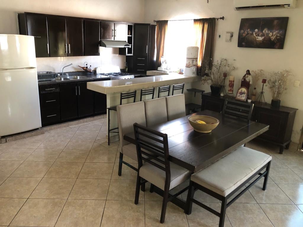 An open kitchen and dining area is displayed, featuring dark wooden cabinets and a light countertop. A dining table with soft seating accommodates several guests. Natural light enters through the window adorned with dark curtains, enhancing the relaxed atmosphere.