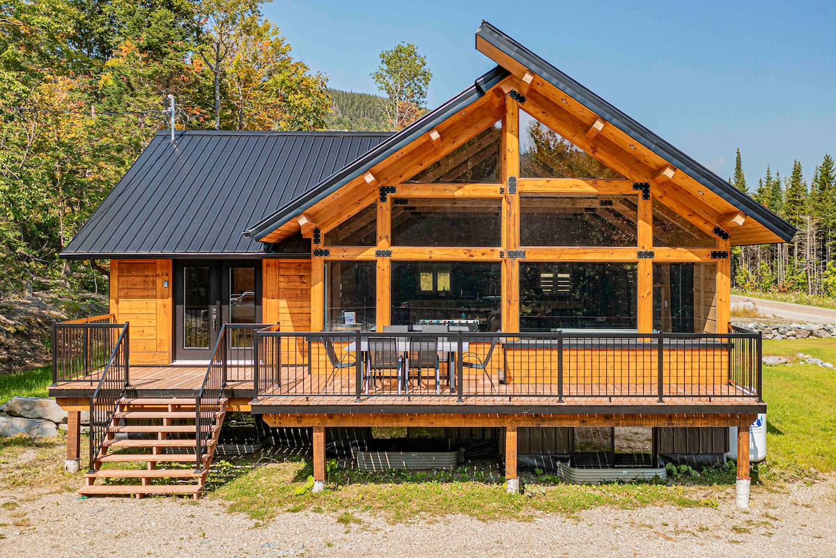 The exterior of a modern rustic cabin features a large wooden deck with a railing, several outdoor chairs arranged around a table, and expansive glass windows that invite natural light. The cabin is surrounded by greenery and distant mountains, creating a serene setting.