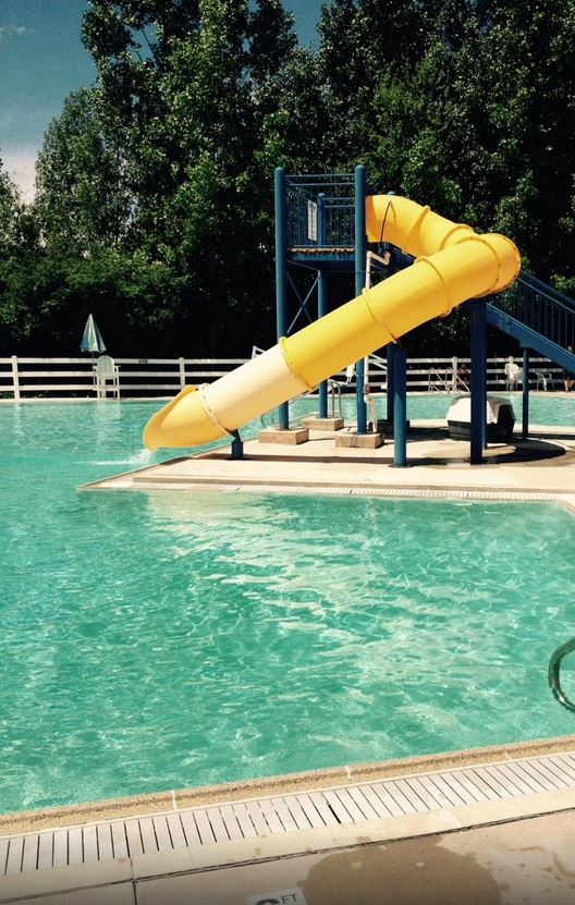 A clear blue swimming pool features a vibrant yellow water slide, with shaded loungers positioned nearby. Lush green trees provide a natural backdrop, enhancing the recreational area. The pool area is well-maintained, inviting guests to relax and enjoy the outdoor ambiance.