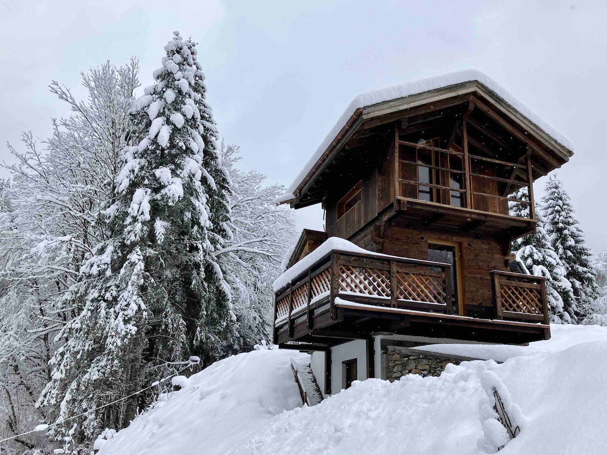 The cozy chalet features a traditional design, elevated on a snow-covered landscape. It is surrounded by tall evergreen trees, with a spacious balcony overlooking the serene winter scenery. Soft snow blankets the ground, creating a peaceful atmosphere.