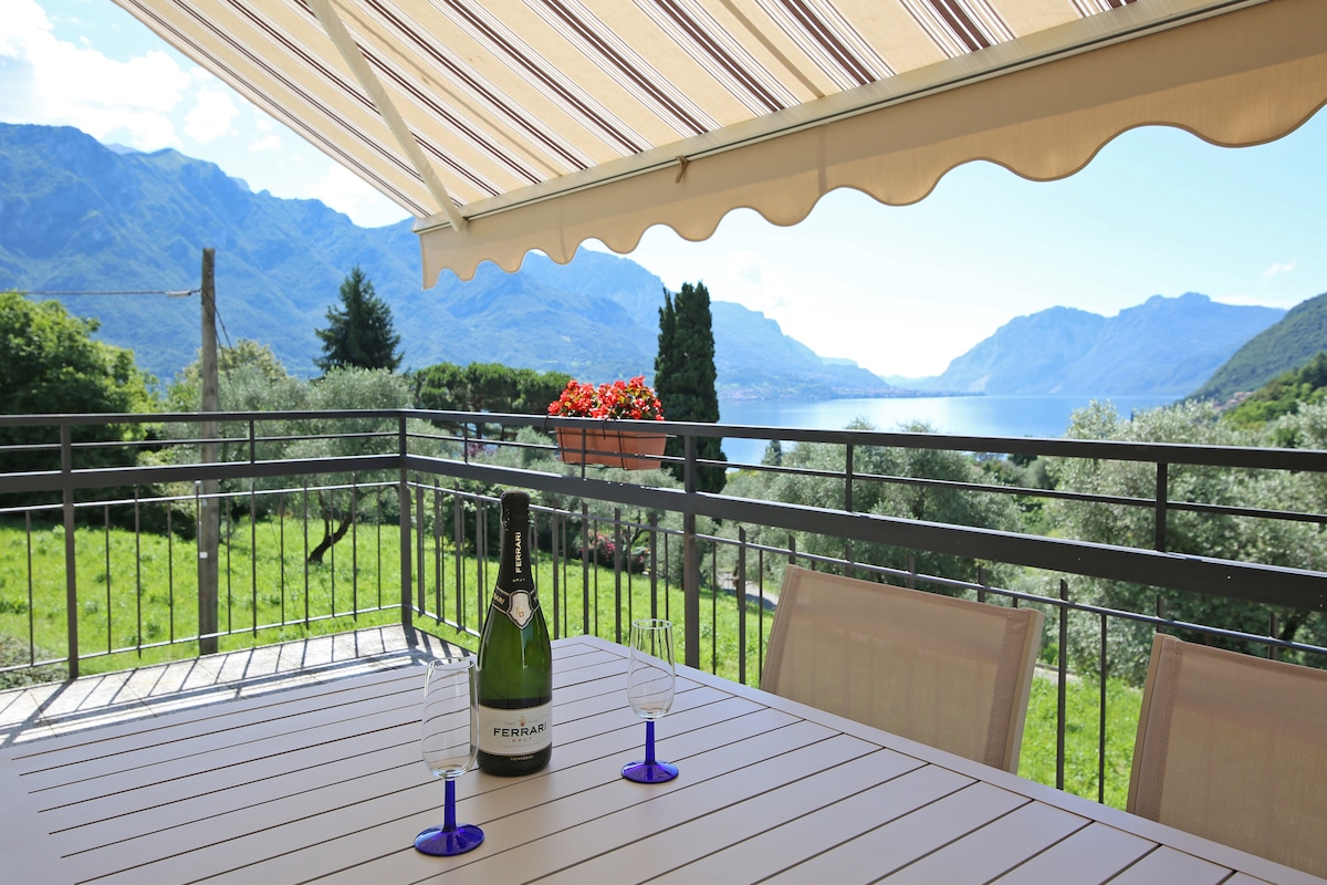 An outdoor terrace is furnished with a dining table and chairs, offering a view of the lake and surrounding mountains. A bottle of sparkling wine and two glasses are set on the table, inviting guests to enjoy the scenic landscape under an awning.
