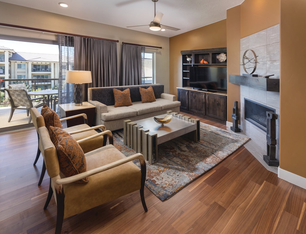 A spacious living area is presented, featuring a comfortable sectional sofa paired with accent chairs. A wooden coffee table sits atop a decorative rug, while a wall-mounted television is positioned above a stylish fireplace. Natural light enters through large windows framed with dark curtains.