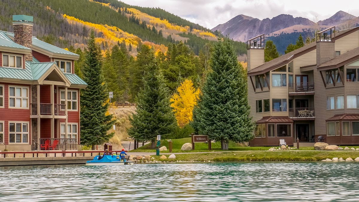 Two multi-story condominium buildings are visible along the shoreline of a calm lake, surrounded by vibrant autumn foliage. The red building features a stone chimney and red accents, while the other presents a more neutral color palette. A small blue watercraft is docked nearby.