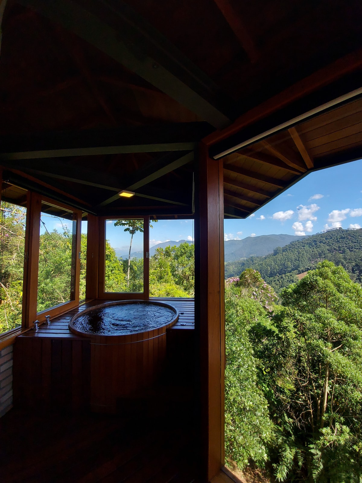 A wooden hot tub is positioned on a covered deck, offering expansive views of lush mountains and a clear blue sky. Large windows frame the scene, inviting natural light and a sense of tranquility into the space.