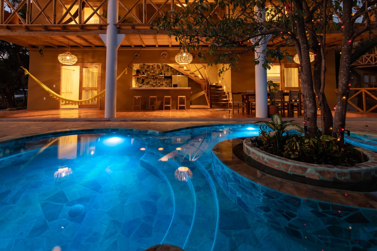 A private pool is featured with a glimmering blue surface, surrounded by natural stone accents. The area includes lush greenery and is illuminated by soft lighting. The villa’s open space is visible in the background, showcasing a stylish bar and dining area.
