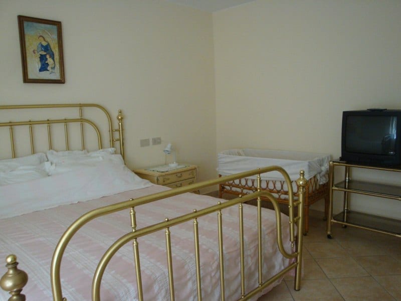 A warmly lit bedroom features a gold metal bed frame with a spacious bed. A small crib is positioned beside it. A simple side table with a lamp is nearby, and a television is placed on a metal stand. Light walls enhance the room's spacious feel.