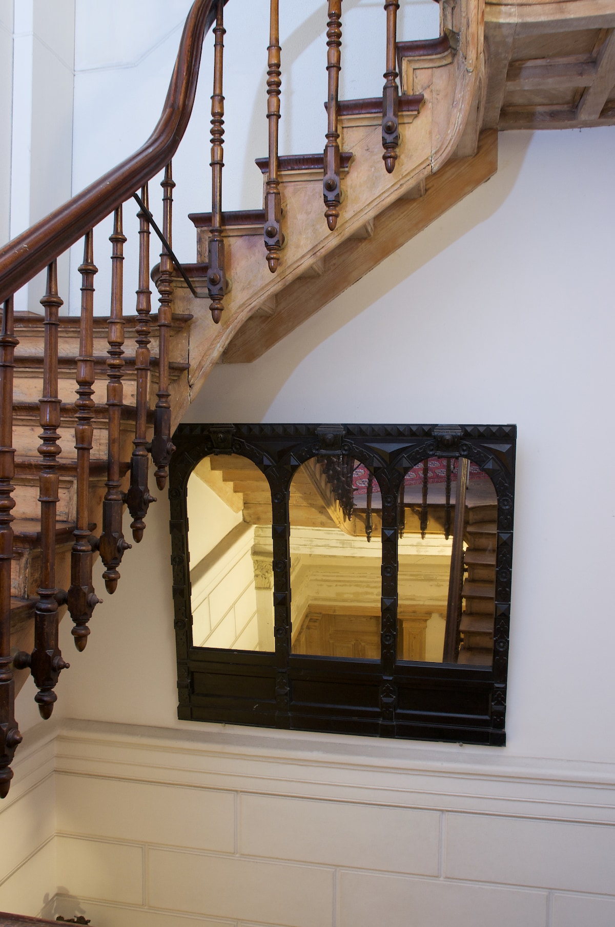 A staircase constructed of polished wood ascends to the upper floors. An ornate black-framed mirror, featuring arched detailing, is mounted on the wall adjacent to the stairs, reflecting the elegant design and charm of the historic building.
