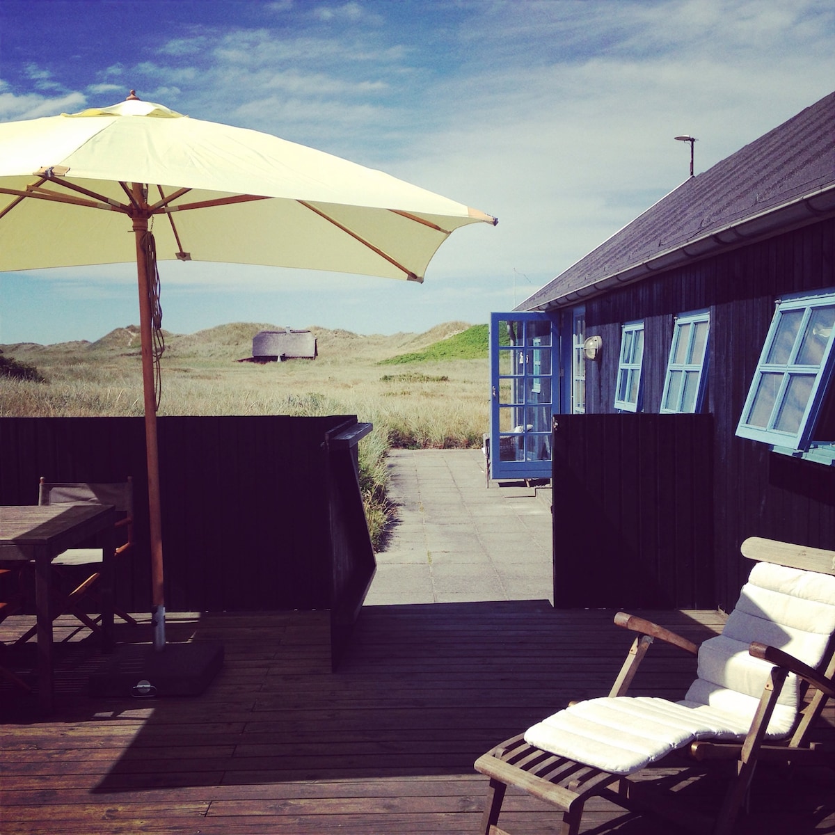 A sun umbrella provides shade over a wooden deck area furnished with seating. Blue-framed windows line the black exterior of the house, which leads to a pathway through the grassy dunes. The sky is clear, contributing to a bright atmosphere.