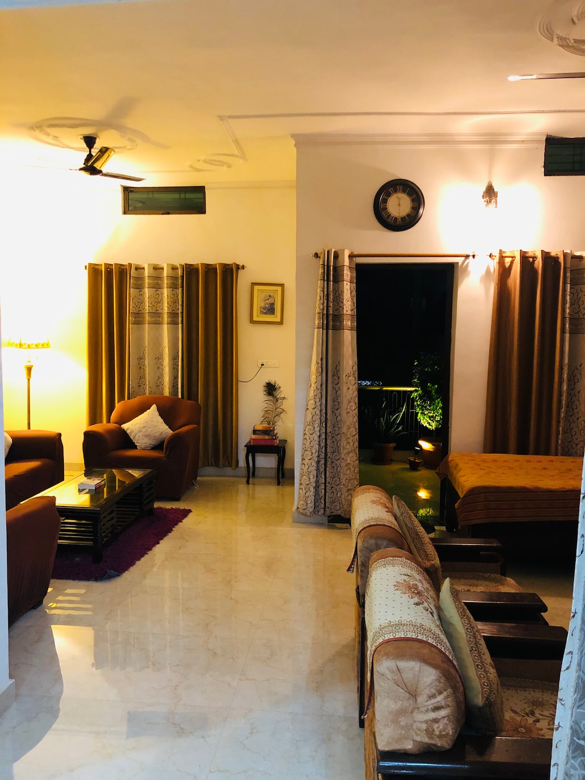 The spacious lounge area is furnished with comfortable seating arrangements, featuring two armchairs, a sofa, and a side table. Natural light filters through the window drapes, and a lamp provides warm illumination. A balcony view with greenery adds a serene touch to the ambiance.