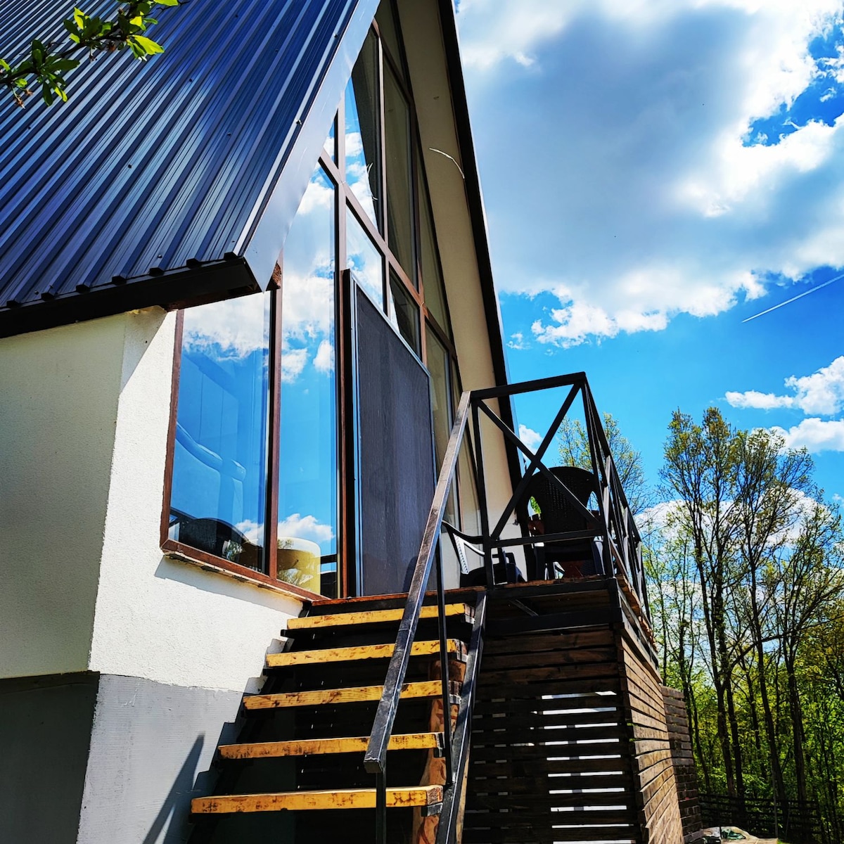 A modern house is depicted with large windows reflecting the sky, highlighting the architectural design. A wooden stairway leads to the entrance, surrounded by lush greenery and bright blue skies with scattered clouds.