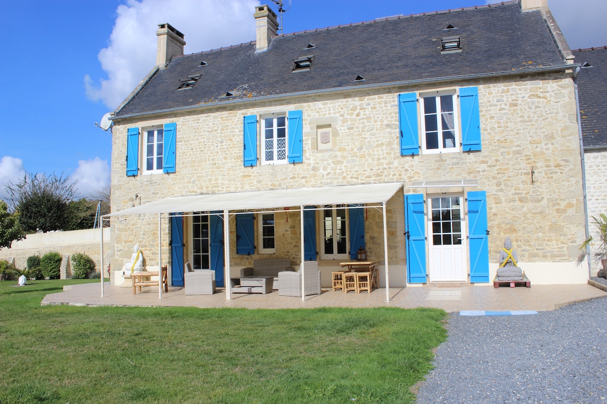 The exterior of a stone house is showcased, featuring a covered patio area furnished with seating and a dining table. Bright blue shutters frame the windows, complementing the beige stone walls. A landscaped lawn extends in front of the house, creating an inviting outdoor space.