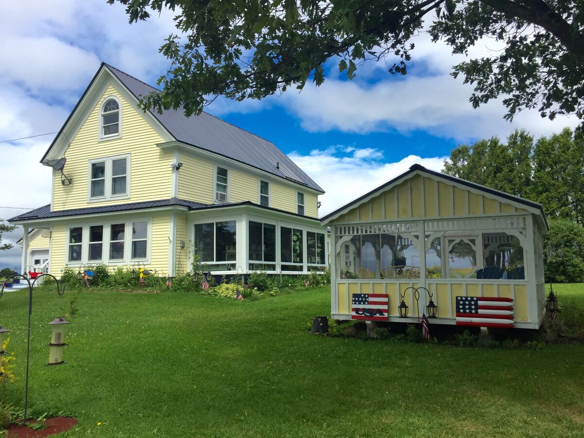 A charming yellow farmhouse with a gray roof is set amidst a lush green lawn. A gazebo with American flags on its side complements the house, while a blue sky and scattered clouds provide a serene backdrop.