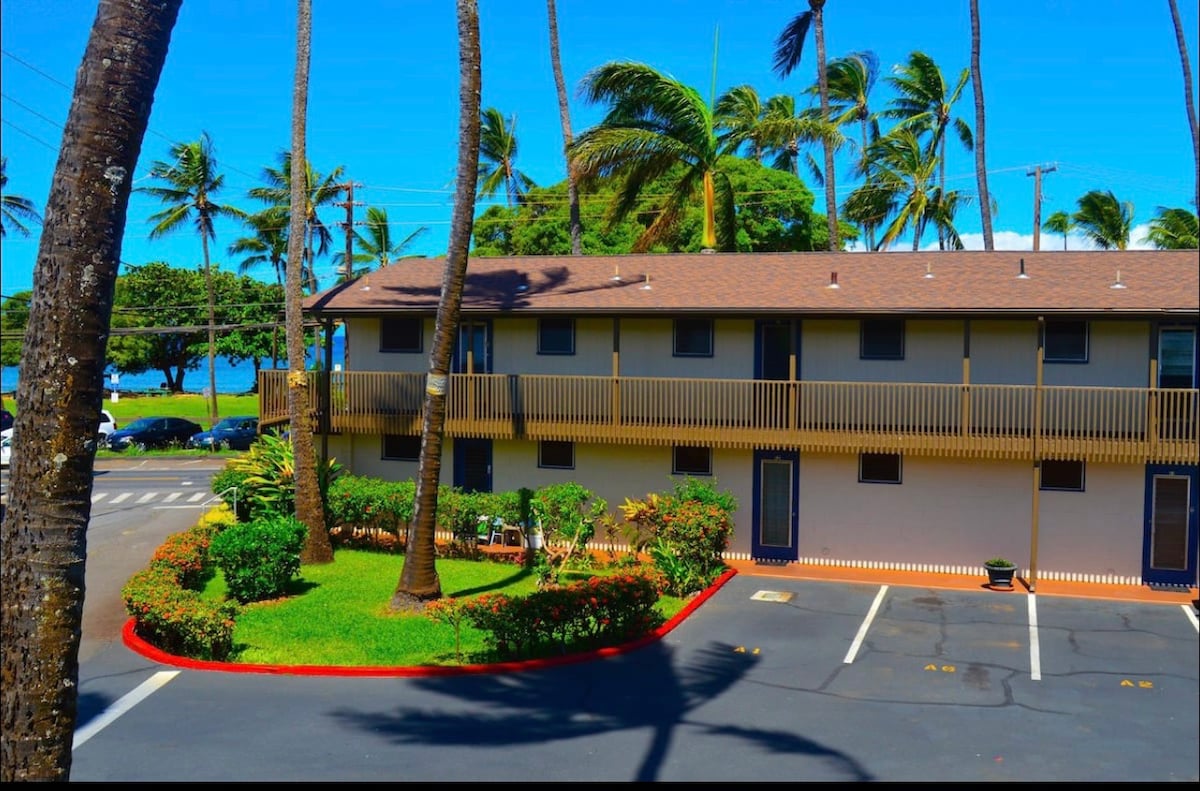 A two-story building is surrounded by palm trees and lush greenery, with a clear blue sky overhead. The exterior features light-colored walls and a wraparound balcony. A well-maintained parking area is visible in front, along with a glimpse of the ocean in the background.