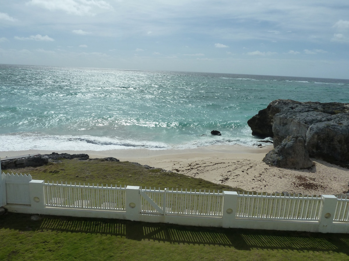 A tranquil beach scene is viewed from the villa, featuring gentle waves lapping at the sandy shore. The ocean glimmers under the sunlight, while a low rocky outcrop is visible to the right. A white picket fence borders the grassy area, adding charm to the coastal setting.
