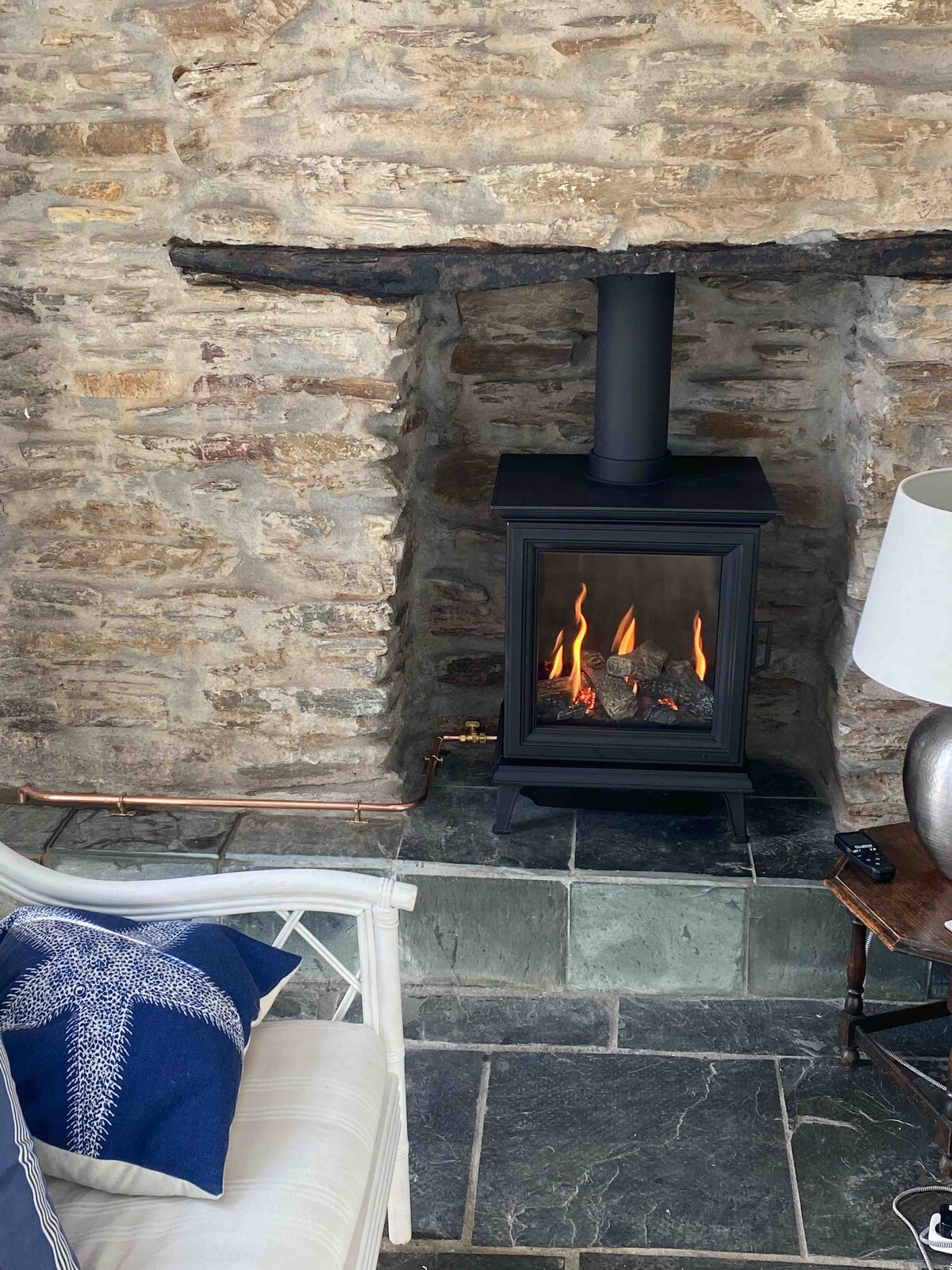 A modern wood-burning stove is set against a rustic stone wall, creating a cozy focal point. The stove emits a warm glow, with flames visible through the glass front. A comfortable chair with a nautical-themed cushion is nearby, accompanied by a side table and lamp.