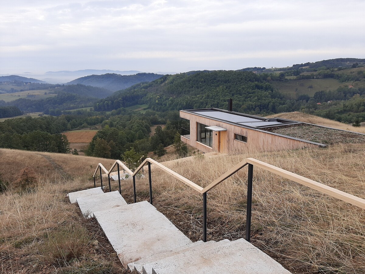 A modern house is situated on a hill, featuring a green roof and large windows. A set of stairs leads down from the house, surrounded by tall grasses and a panoramic view of rolling hills and valleys in the background.