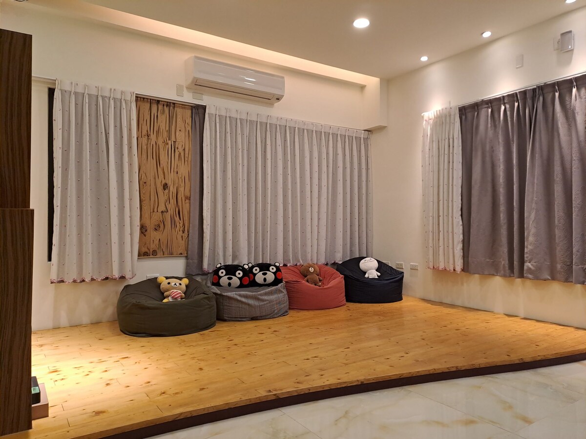 A warm, wooden-floored space features several plush bean bags in varied colors, positioned against soft draped curtains. Natural light filters in through the windows, complementing the inviting ambiance of the area.