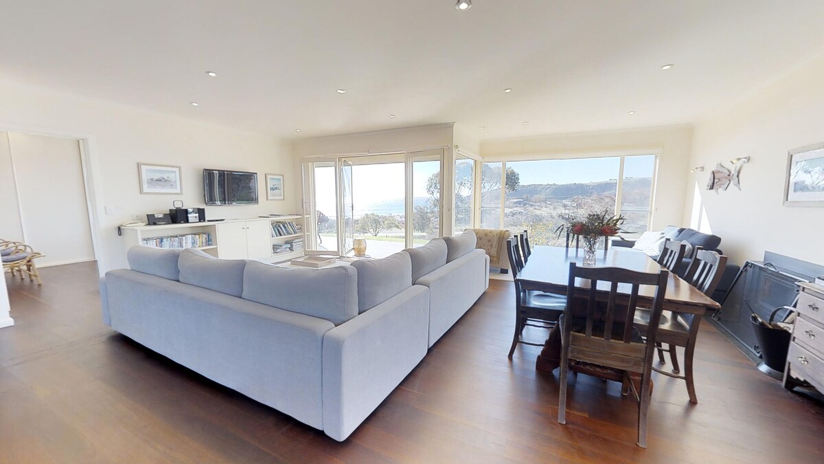 A spacious living area is shown, featuring a light-colored sectional sofa and a wooden dining table surrounded by chairs. Large windows provide a view of the outdoors, allowing natural light to fill the space. A TV unit is situated in the corner.