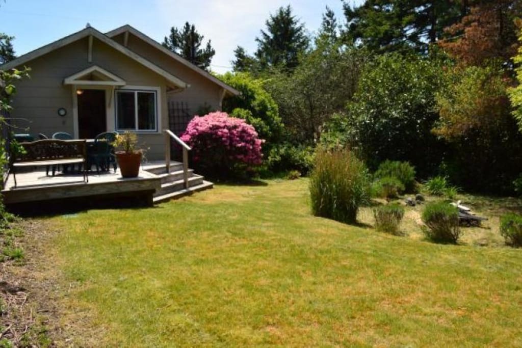 Large grassy landscaped backyard with firepit and deck with table, chairs and charcoal BBQ.