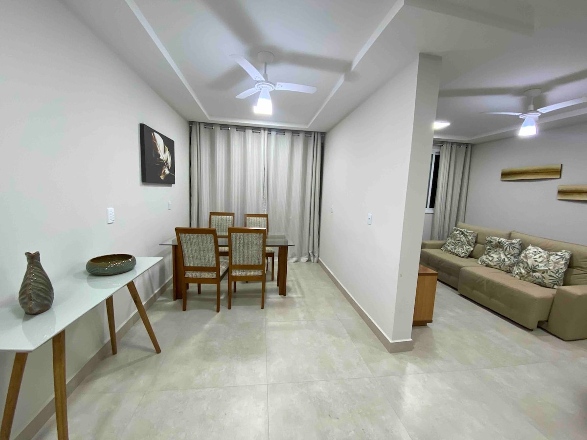 A modern living area is presented, featuring a light-colored sofa and a dining table with four chairs. Soft lighting creates a relaxed atmosphere, while light-colored walls and a ceiling fan enhance air circulation. Decorative elements include a wall art piece and a small decorative vase.