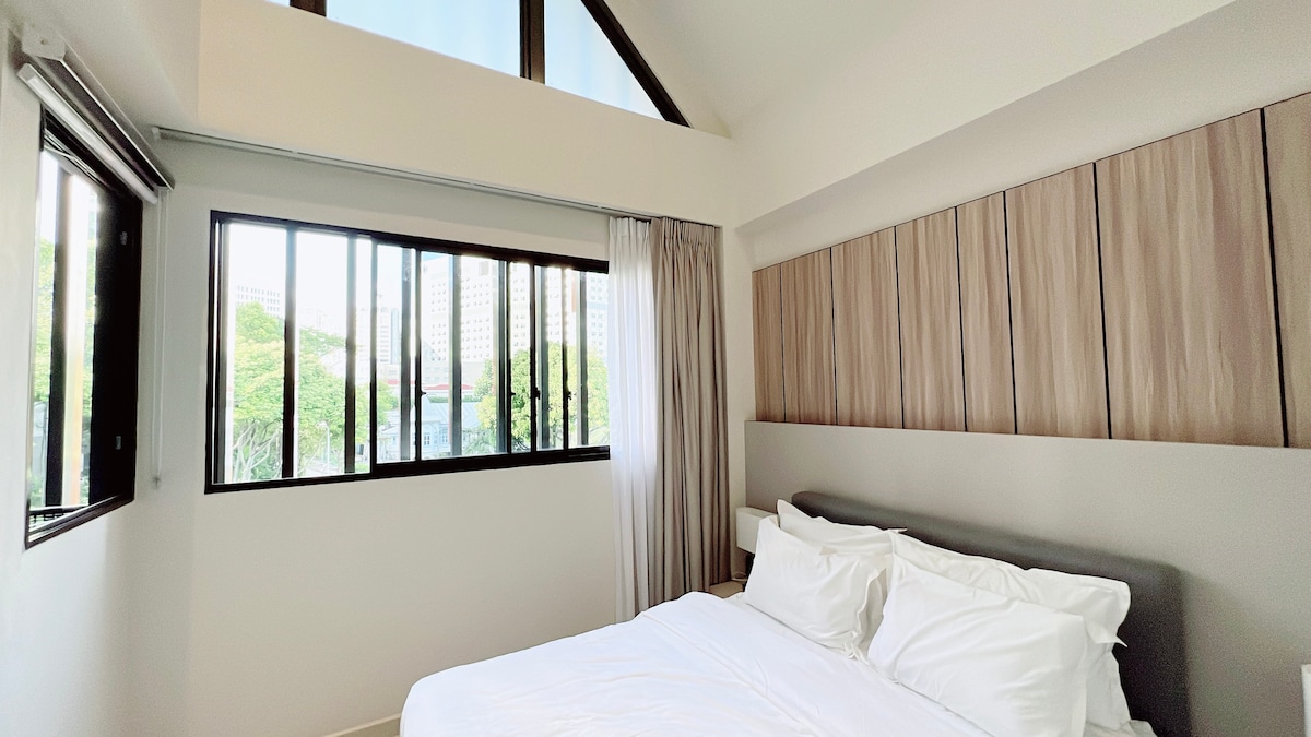 A bright bedroom features a spacious bed with crisp white linens against a modern wooden headboard. Large windows with black frames allow natural light to fill the space, complemented by high ceilings and soft drapes that create a serene environment.