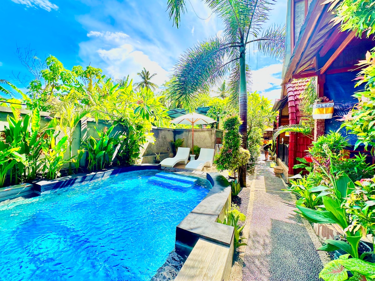 A seasonal outdoor swimming pool is surrounded by lush tropical plants, with lounge chairs positioned for relaxation. The area is brightened by sunlight filtering through palm trees, and a shaded umbrella provides an inviting spot for rest beside the water.