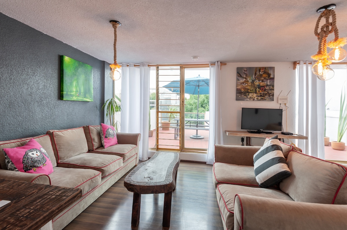 A comfortable living area is furnished with two sizable couches featuring pink and striped accents. Natural light filters through curtains, illuminating a rustic wooden coffee table. A media console with a TV is positioned next to a sliding glass door that leads to an inviting terrace with an umbrella.