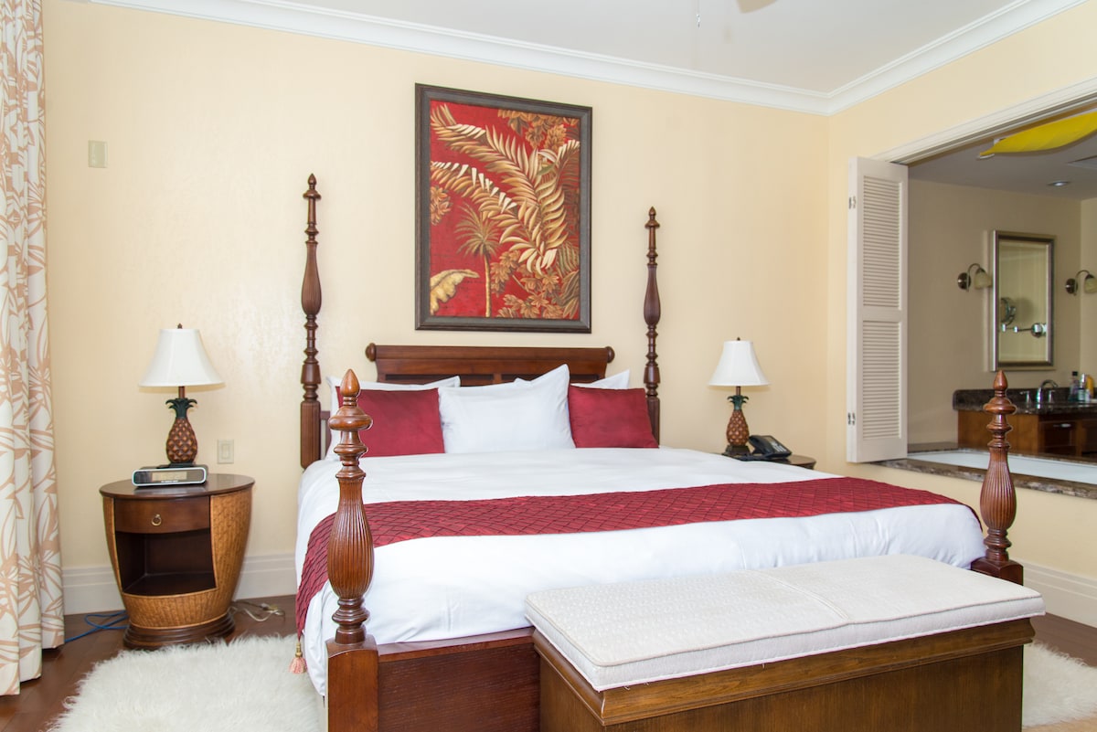 A spacious bedroom featuring a large king-size bed with decorative red and white bedding. Flanking the bed are two bedside lamps on wooden tables. A bench is placed at the foot of the bed. A lush botanical artwork adorns the wall above the bed, complementing the neutral-toned decor.
