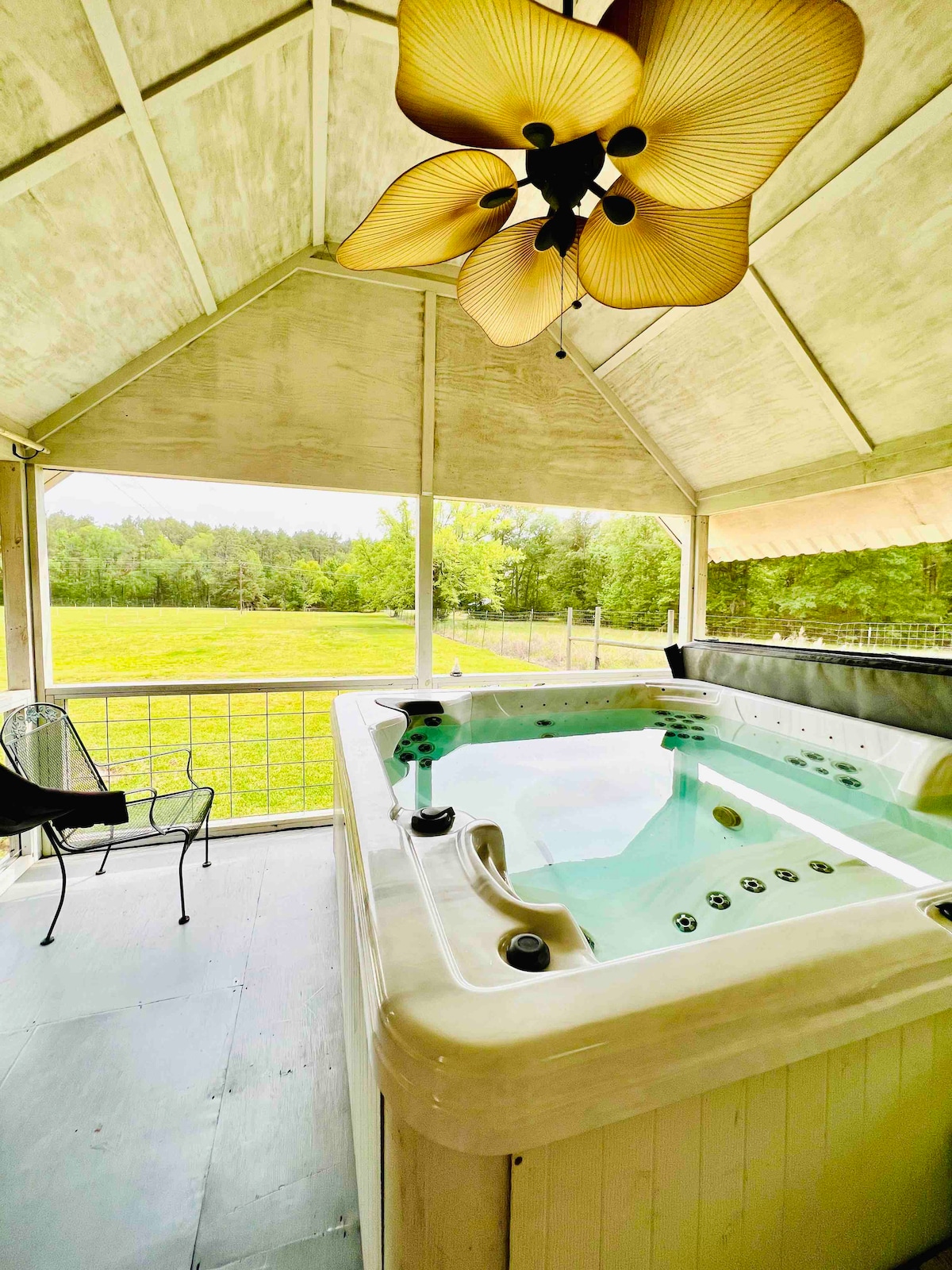 A spacious hot tub is situated under a high, open canopy, surrounded by verdant greenery. Nearby, a black chair offers a place to relax. Soft natural light illuminates the area, creating a serene environment with views of the expansive lawn beyond.