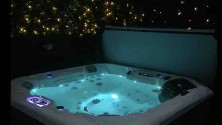 A modern hot tub is enveloped by a serene nighttime atmosphere, showcasing soothing blue water. Warm light from built-in jets complements the sparkling ambiance created by twinkling fairy lights in the background, enhancing the space's overall tranquility.