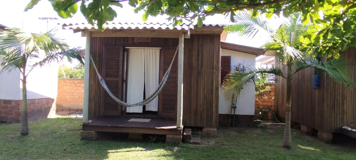 A rustic wooden cabin is surrounded by lush greenery, featuring a small porch with a hammock. Light filters through the trees, creating a serene outdoor space. The structure is paired with a larger building nearby, offering space for relaxation.