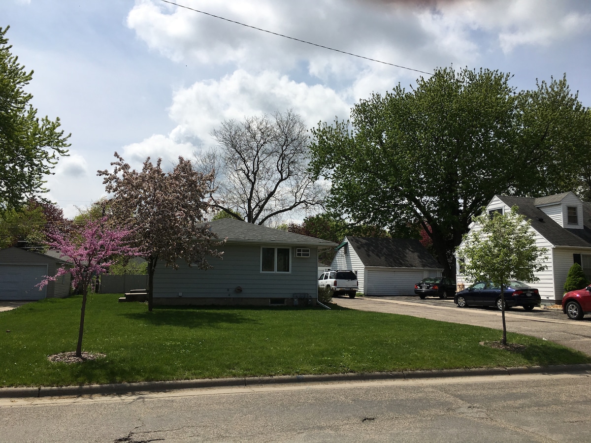 A well-maintained duplex features a grassy front yard, flanked by blooming trees. The structure displays a neutral exterior with plenty of natural light. Nearby, parked vehicles can be seen along the driveway, featuring a mix of trees and greenery in the background.