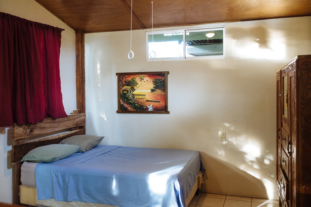 A cozy sleeping area is designed with a wooden headboard, and a comfortable bed covered with a blue blanket. Natural light enters through a small window, illuminating the room, which features an art piece on the wall and red curtains that add a touch of color.