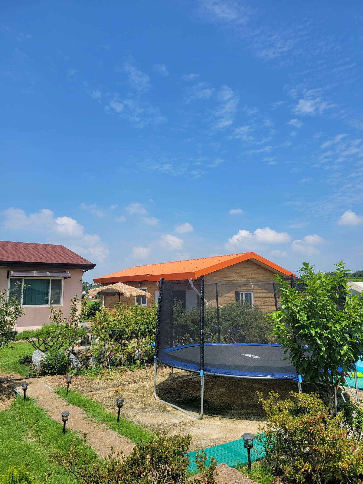 A spacious garden area features a trampoline situated beside a wooden cabin with a vibrant orange roof. Lush greenery surrounds the trampoline, and a clear blue sky completes the tranquil outdoor scene.