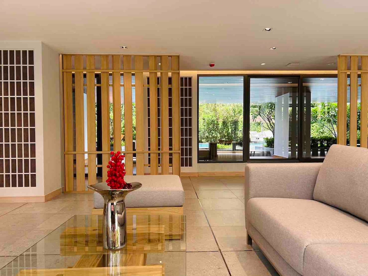 A minimalist lobby area is presented, featuring a light-colored sofa and a glass coffee table with a silver vase holding vibrant red flowers. Large windows provide a view of the greenery outside, enhancing the space's open feel.