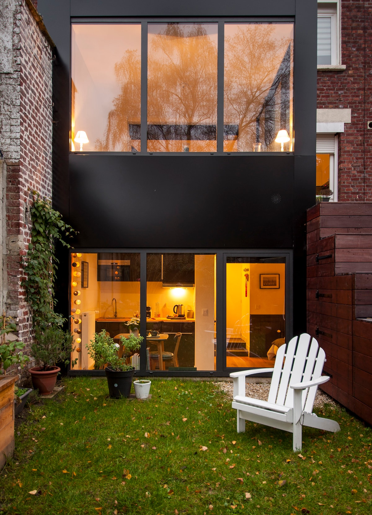 A modern garden area is visible, featuring a white lounge chair positioned on green grass. The back of a studio showcases large glass windows illuminated from within, revealing a cozy interior space. Potted plants add a touch of greenery to the scene.