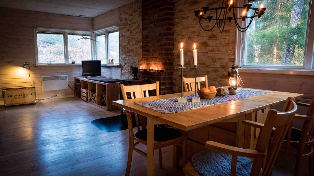 An inviting dining area is highlighted by a wooden table surrounded by several light-colored chairs. Natural light filters through large windows, illuminating the space. A cozy sitting area features a sofa and a television, while soft candlelight adds warmth to the interior.