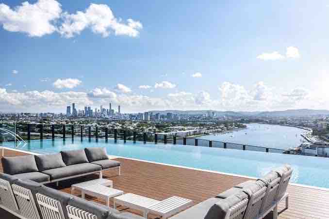 A rooftop pool area is showcased with modern lounge seating arranged on wooden decking. The expansive view includes the Brisbane skyline and the river below, complemented by clear blue skies and scattered clouds.