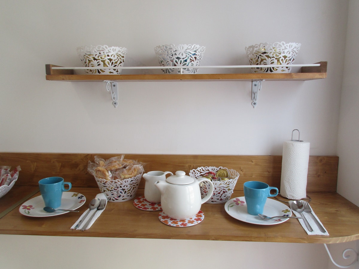 A wooden shelf displays intricately designed white bowls, alongside a teapot and two turquoise cups. Placemats, silverware, and a stack of pastries are arranged on a wooden surface, creating an inviting setting for breakfast. A paper towel holder is mounted nearby.