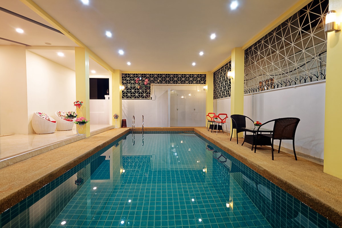 A shared indoor swimming pool is featured, surrounded by a smooth tiled surface. Comfortable seating arrangements are positioned nearby, including two black chairs and a small table. Soft lighting highlights the relaxing ambiance, while potted flowers add a touch of color to the space.