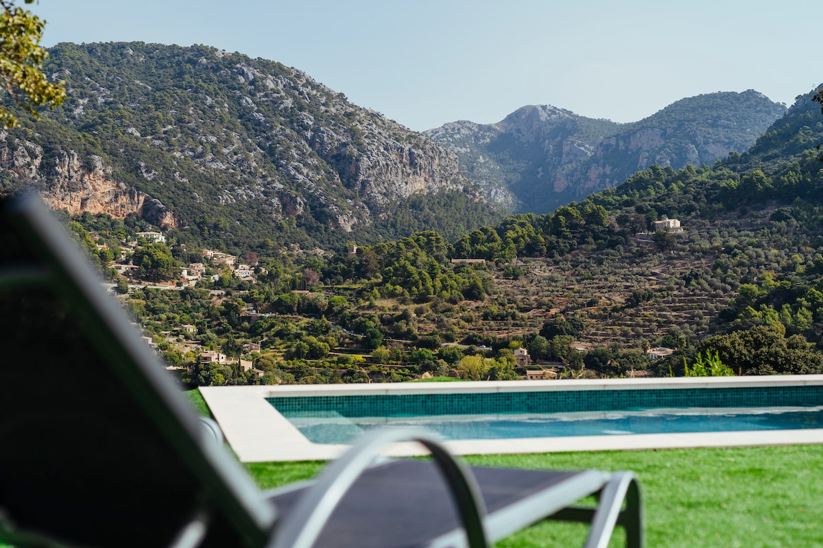 A swimming pool reflects the clear blue sky, with lounging chairs positioned nearby. The surrounding landscape features rolling green hills and majestic mountains, providing a serene backdrop to the outdoor space. Lush vegetation and terraced fields are visible in the distance.