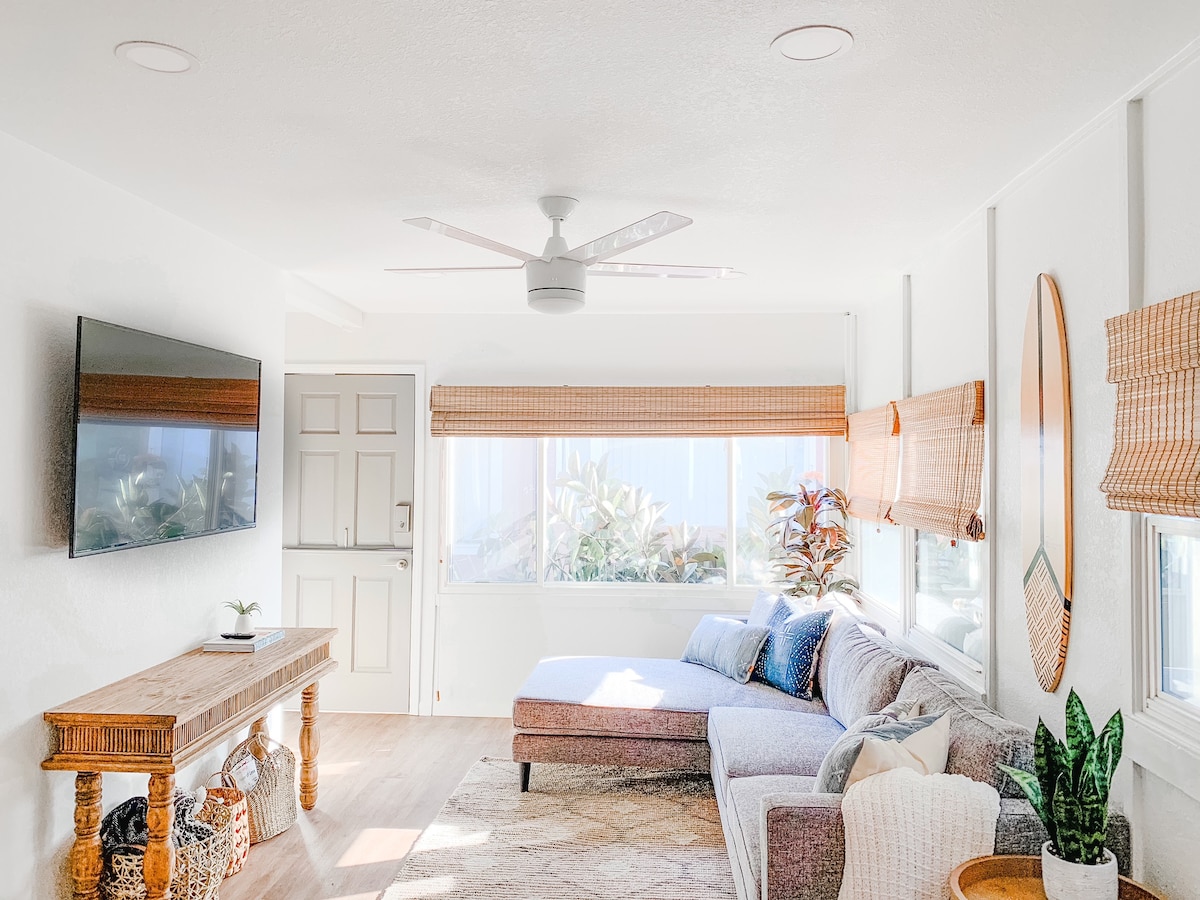 A bright living room features large windows that invite natural light. A comfortable sectional sofa is positioned next to a wooden table. A wall-mounted smart TV is visible across from the seating area. Indoor plants add a touch of greenery to the space.