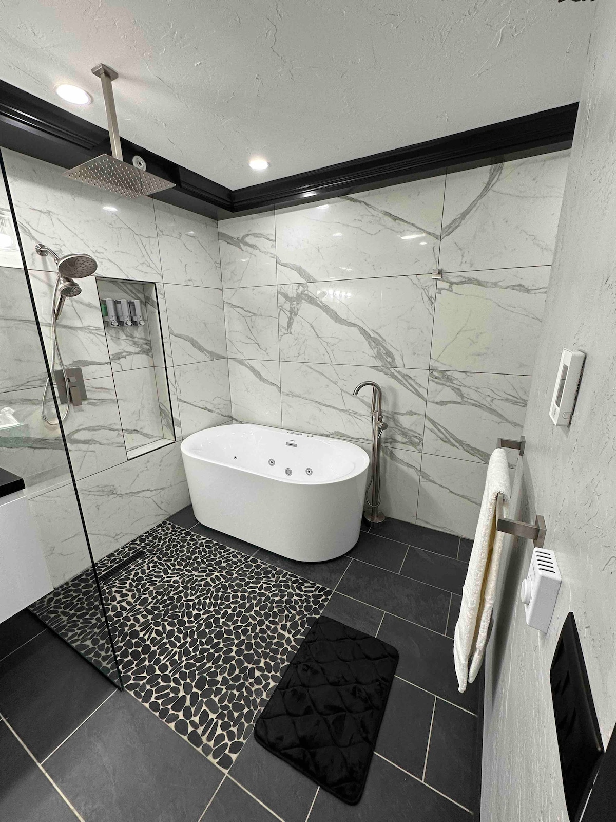 A modern bathroom features sleek marble walls and a freestanding white tub. A glass shower enclosure is visible alongside a dark stone floor. A plush black mat and a white towel are neatly positioned near the tub, contributing to the overall clean and contemporary design.