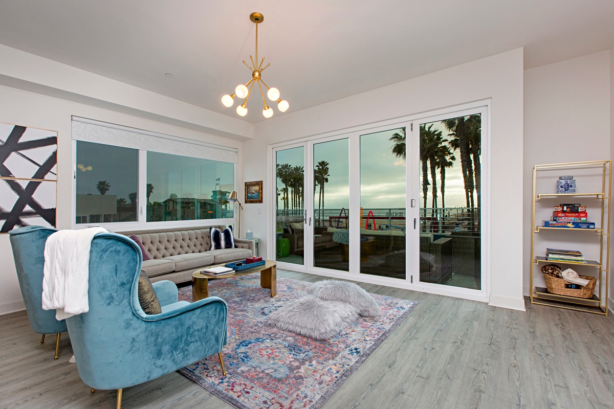A bright living area features large glass doors that open to a palm-lined view outside. A light blue velvet armchair and a beige tufted sofa complement a decorative area rug. A modern chandelier hangs from the ceiling, adding a touch of elegance.