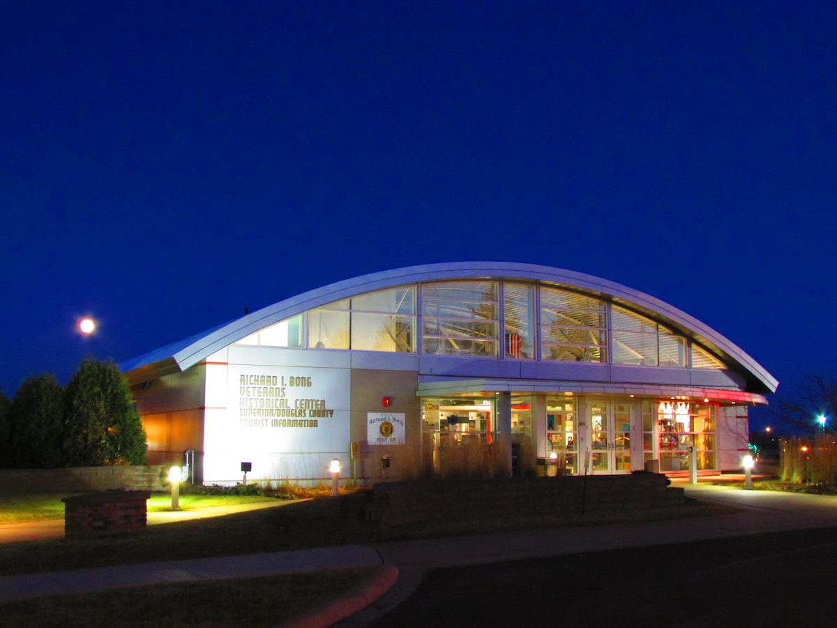 The building features a modern architectural design with a curved roof and large glass panels. Soft lighting illuminates the entrance, creating a welcoming atmosphere. The setting showcases a clear night sky with a prominent moon, enhancing the evening ambiance.