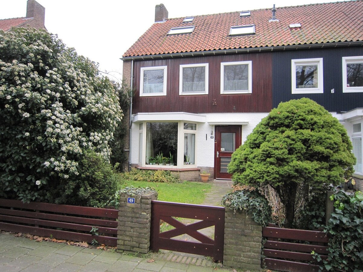 The exterior of a two-story house is presented, featuring a combination of wood and brick materials. Lush greenery surrounds the entrance, with a pathway leading to the front door. Large windows provide a view of the interior, framed by flowering bushes.