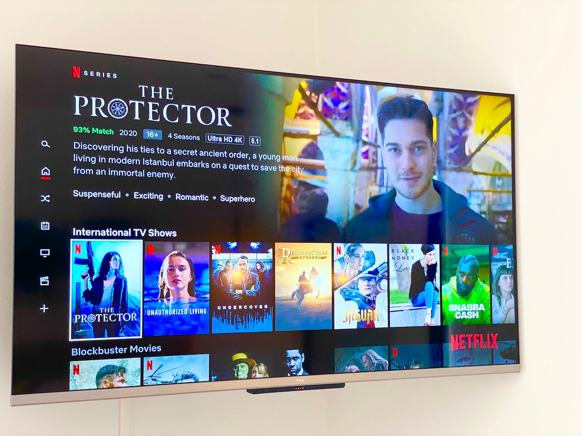 A flat-screen television displays the Netflix homepage, featuring dynamic thumbnails of various TV shows and movies. The interface highlights the series 'The Protector,' suggesting options for viewing and exploring content.