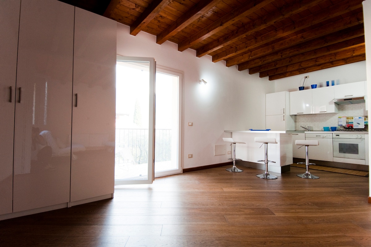 A bright kitchen area is shown with modern white cabinetry and a bar-style table featuring three stools. Large glass doors open to a balcony, allowing natural light to flood the space. Wooden beams provide a warm contrast to the sleek surfaces.