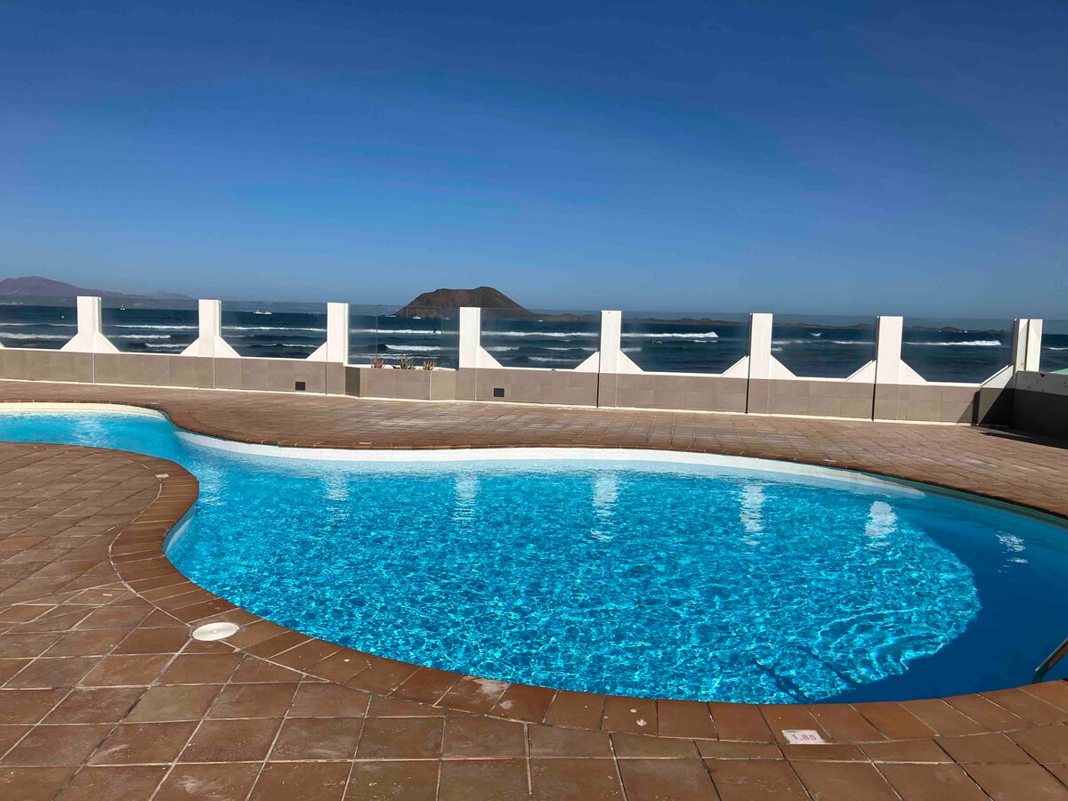 A clear blue swimming pool is surrounded by tiled decking and offers panoramic views of the ocean and nearby island. The horizon showcases gentle waves under a bright sky, creating a serene outdoor atmosphere.
